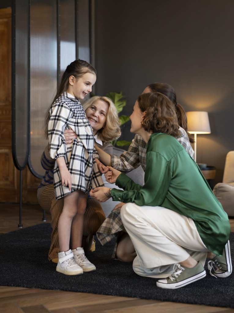 A young girl in a plaid shirt dress standing while a woman in a green blouse and an older woman kneel beside her, smiling and holding her hands.
